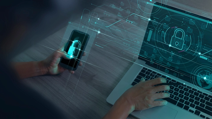 hand holding iphone and another hand typing on open laptop with cybersecurity lock symbols