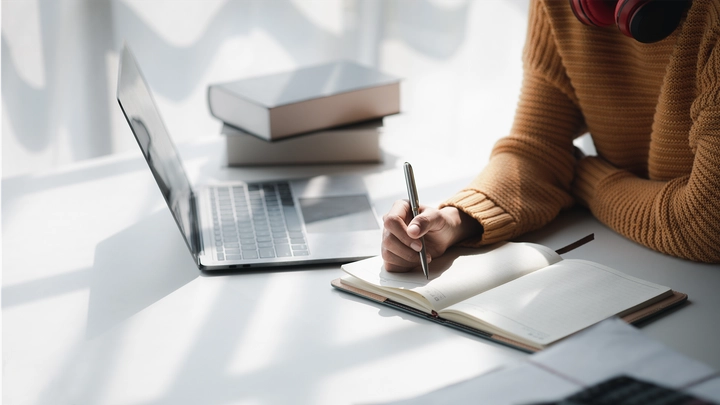 white wall and desk with hands taking notes next to laptop